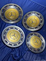 Four Mercedes Benz gold and chrome hub caps laid on dark blue fabric, showing front side with emblem and rims.