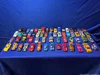 Two photos showing 60 small die-cast toy cars including Matchbox and Hot Wheels models arranged on a dark surface with blue backdrop. Cars vary in colors and styles including sports cars, trucks, vintage models and fantasy designs.