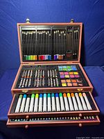 Wooden art kit box open showing multiple compartments with art supplies including colored pencils, markers, watercolor paints, oil pastels, chalk pastels, paintbrushes, pencil sharpener, and eraser.