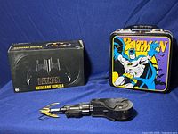 Photo shows Batman lunchbox with handle, Batman Batarang replica box, and Bat rappel replica toy on blue background.
