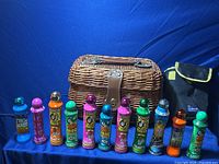 Ten colorful bingo dabbers and a rectangular brown wicker basket with leather strap and metal clasp, arranged in front of a blue background.