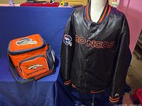 Black Denver Broncos jacket on mannequin next to orange and blue Broncos cooler bag.