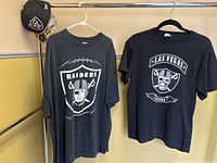 Two black Oakland Raiders themed t-shirts hanging on hangers next to a black baseball cap with a Raiders logo hanging on the side of a cabinet.
