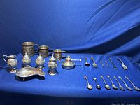 Full display of assorted metal collectible items on blue fabric background including cups, mugs, small containers, spoons, forks, and a sauce dish.