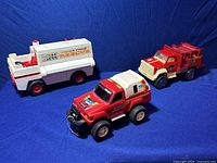 Photo of all three toy vehicles together on a blue background: white Fisher Price rescue ambulance with red accents and two red fire trucks.