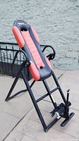 Photo showing the Health Gear Teter inversion table, focusing on the red and black padded seat, armrests, and foot holding mechanism, against an outdoor background.