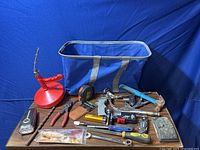 Various hand tools arranged on a table with a large blue tool bag behind them