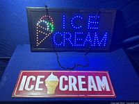 Two ice cream signs in a dark setting: an electric LED lighted sign showing colorful lights forming an ice cream cone and the words ICE CREAM, and a metal sign with red background, white text, and ice cream cone graphic.