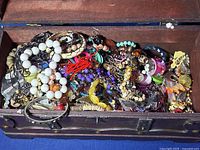 Close-up of miscellaneous colorful jewelry pieces tangled together inside a wooden chest, showing beads, rings, bracelets, and earrings.