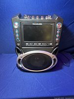 Front view of Karaoke USA karaoke machine featuring a 7-inch LCD screen, multiple control buttons, and a large built-in speaker with silver grill.