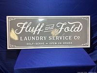 Front-facing photo of the gray metal sign with white lettering reading 'Fluff and Fold Laundry Service Co. Self-Serve Open 24 Hours'.
