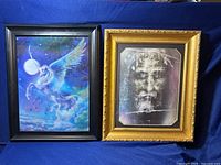 Two framed lenticular artworks displayed side by side on a blue cloth background. Right frame is the grayscale Jesus Christ face with black and gold border. Left frame is colorful Pegasus flying at night scene with black frame.