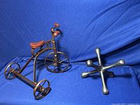 Overview photo showing a metal tricycle bike sculpture with a brown seat and handle grips next to a large metal jack with rounded ends on a blue fabric background.