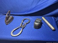 Four metal items on a blue fabric background: a cast iron iron, horseshoe shaped puzzle piece, a cutter sharpener, and birth certificate holder cylindrical tube.