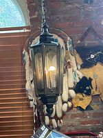 Mid century swag hanging lamp with hexagonal metal frame and perforated panels, illuminated bulb. Lamp hangs by a long metal chain in front of a brick wall and window with wooden blinds. A textile dreamcatcher or wall hanging is visible in the background.