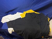 Photo of all eleven youth t-shirts in black, yellow, and white colors, folded and stacked on a blue surface.