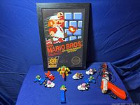 Wide view of the lot showing the framed Super Mario Bros. cover art, multiple small Mario Bros. character toys (with figures riding motorbikes), Bowser figure, and the original Duck Hunt Zapper light gun with its cord.