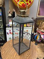 Front and angled view of the large outdoor candle holder lantern showing the black metal frame, clear glass sides, and brass-colored top with handle.