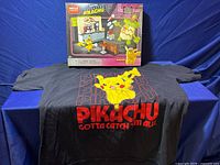 Black Pikachu T-shirt with large graphic and text, laid flat beneath boxed Mega Construx Detective Pikachu game board set