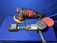 Two power tools: red and black car polisher with buffing pad, black and gray cordless angle cutter with cutting disc and 20V battery pack on a blue cloth.
