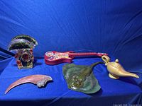 Four novelty items displayed on blue cloth background: glass mushroom napkin holder with red accents, red toy guitar, green stingray figurine, gold genie lamp, curved claw-shaped object