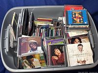 Large plastic bin filled with assorted CD cases featuring a mix of artist images and case styles