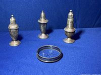 Three sterling silver weighted salt and pepper shakers with varying designs tarnished with age, standing upright on a blue background along with a small sterling silver & crystal ashtray or cup holder with pierced gallery edge and cut crystal base.