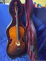 Open cello case displaying the student cello inside with natural brown wood finish, four strings, and black fingerboard.