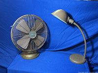 Photo showing vintage brown desk lamp and vintage repro style brown desk fan side by side on blue background.