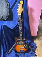Full view of vintage Harmony sunburst electric guitar standing on a guitar stand against a blue backdrop.