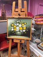 Painting showing white and yellow roses in a green vase, mounted on an easel with a gold frame.