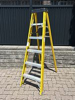 Yellow 6-foot Reynolds fiberglass step ladder standing open on outdoor paving with a black metal fence and stone wall background. The ladder has six flat aluminum steps and no paint shelf on the top cap.