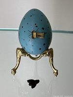 Front view of blue egg measuring tape on gold-tone tripod stand with detached leather bee in foreground