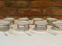 Front view of eight porcelain ramekins seated in pierced sterling holders with spoons arranged in a row