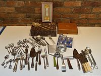 Full overhead view of mixed flatware and serving utensils laid out on white table with brick wall background.