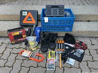 Photo showing full lot including CAA emergency kit, jumper cables, traction mat in box, Mercedes Benz truck box, black shoe covers, assorted tools, and emergency supplies all arranged on pavement.