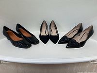 Three pairs of black Ron White heels displayed side by side on white surface: woven leather pumps, suede kitten-heel pumps, smooth leather pumps with braided detail