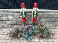 Photo showing two large painted wooden nutcracker figures standing on pavement in front of a stone wall, with three hanging baskets of artificial pine needles, pinecones, and red berries placed at their feet.