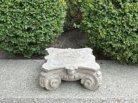 Front view of concrete pedestal on concrete walkway against shrubs showing scrollwork and square top
