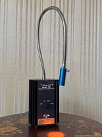 Front view showing device upright, flexible gooseneck, black metal body, mounted on a brown surface with wallpaper background.