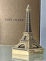 Gold-tone Eiffel Tower shaped solid perfume compact with rhinestone detail on base, reflecting on glass surface against Estée Lauder gold textured box backdrop.