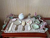 Photo showing various vintage floral-themed vanity items arranged on a wicker and metal tray with floral detail on handles, including brushes, mirrors, porcelain and glass items.