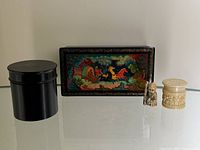 Photo showing all four items: lacquered hand painted trinket box, ebony cylindrical box with lid, and two carved bone figurines on a reflective surface.