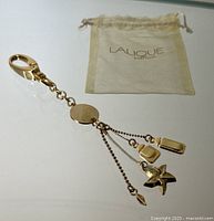 Full view of gold toned Lalique perfume charm keychain with three dangling charms and accompanying branded sheer pouch.