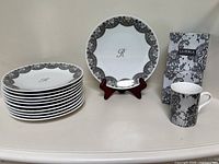 Stack of eleven porcelain dinner plates with black lace Femme design next to La Perla Parfum coffee mugs in box and one mug out of box