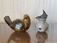 Both ceramic figurines shown side by side on reflective surface with neutral background