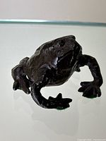 Front angled view of dark brown painted metal frog sculpture with green felt pads on feet showing detailed texture and some paint wear