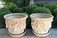 Front view of two round plaster planters with cherub relief and matching saucers on a concrete ledge