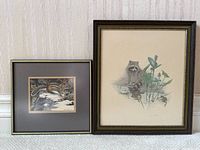 Pair of framed wildlife prints by Robert Bateman, chipmunk and raccoon in dark frames leaning against a wall.