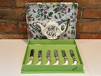 Boxed set displayed open showing six cheese knives with floral ceramic handles and teapot-shaped teabag holder inside box.